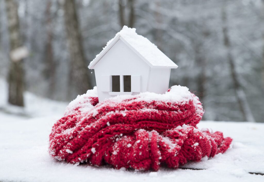 house in winter - heating system concept and cold snowy weather with model of a house wearing a knitted cap